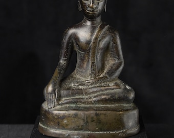 Op de afbeelding: Een bronzen Boeddha-beeld in zittende positie, met een serene uitdrukking. De figuur is gekleed in gewaden en heeft een puntige hoofdtooi. Het beeld staat op een zwarte rechthoekige basis, tegen een donkere achtergrond.