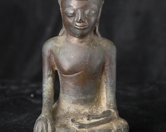 May include: Bronze Buddha statue in a seated meditation pose. The figure has a serene expression, closed eyes, and a pointed headdress. The statue is a weathered bronze color, with visible patina, and sits on a simple base. The background is dark.