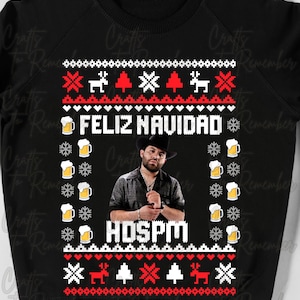 May include: Black Christmas jumper with a red and white snowflake pattern. The jumper features a portrait of a man wearing a cowboy hat and the text "FELIZ NAVIDAD" and "HDSPM".