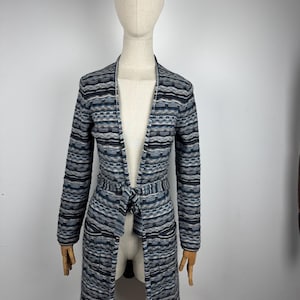 May include: A long, open-front cardigan in a striped pattern of blue, gray, and black. The cardigan features long sleeves, a tie belt at the waist, and two front pockets. The garment is displayed on a mannequin.