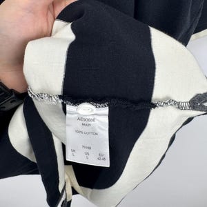 Puede incluir: Camiseta a rayas blancas y negras, 100% algod&oacute;n. La etiqueta indica "AE90686 MULTI 100% COTTON" e informaci&oacute;n de tallas. La camiseta es talla L, talla EU 42-45.