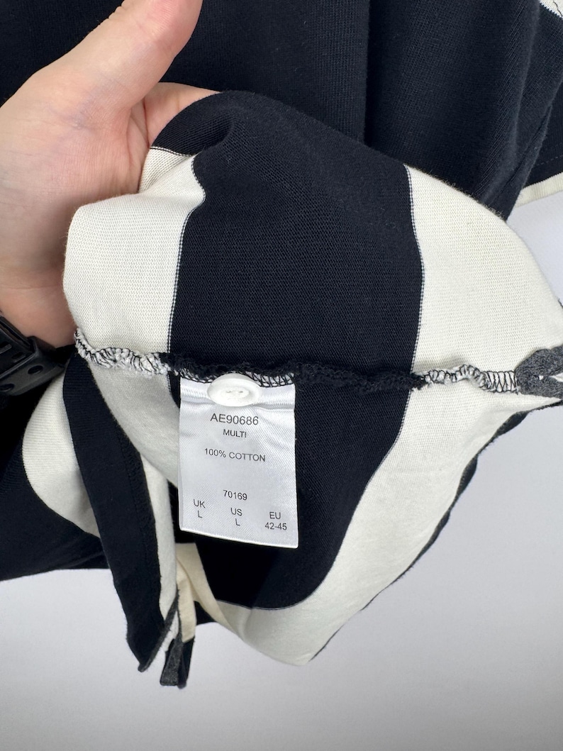 Puede incluir: Camiseta a rayas blancas y negras, 100% algod&oacute;n. La etiqueta indica "AE90686 MULTI 100% COTTON" e informaci&oacute;n de tallas. La camiseta es talla L, talla EU 42-45.