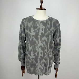 May include: A grey crew neck sweater with a camouflage pattern. The long-sleeved top is displayed on a wooden mannequin against a white background. The sweater's pattern features shades of grey, creating a textured appearance.