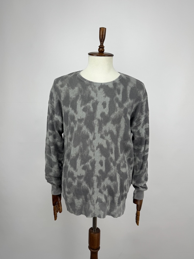 May include: A grey crew neck sweater with a camouflage pattern. The long-sleeved top is displayed on a wooden mannequin against a white background. The sweater's pattern features shades of grey, creating a textured appearance.