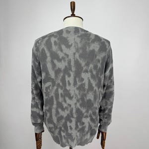 May include: A grey long-sleeved sweater with a textured pattern, displayed on a mannequin. The sweater has a crew neck and ribbed cuffs. The pattern is a lighter shade of grey, creating a camouflage-like design.