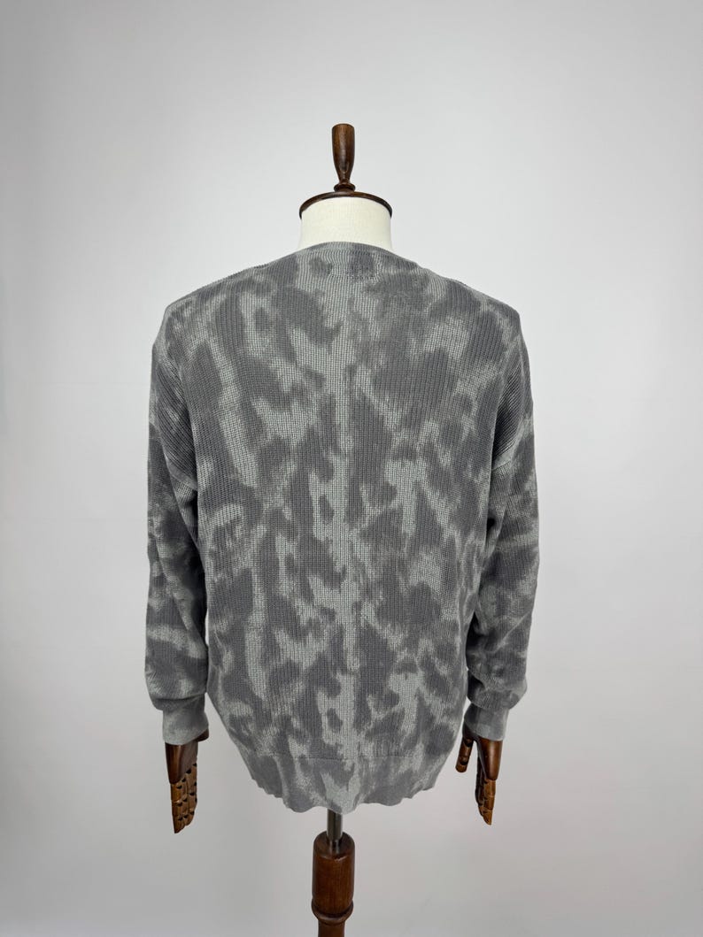 May include: A grey long-sleeved sweater with a textured pattern, displayed on a mannequin. The sweater has a crew neck and ribbed cuffs. The pattern is a lighter shade of grey, creating a camouflage-like design.