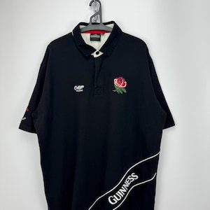 Puede incluir: Polo negro con cuello blanco y una rosa roja bordada en el pecho. La camisa presenta el logotipo de Guinness en blanco en la parte inferior derecha y el logotipo de Cotton Traders en el pecho izquierdo. Ropa deportiva.