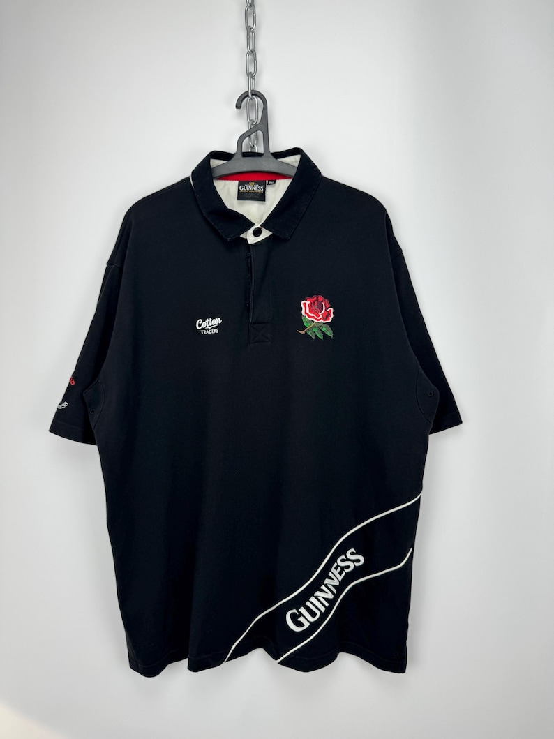 Puede incluir: Polo negro con cuello blanco y una rosa roja bordada en el pecho. La camisa presenta el logotipo de Guinness en blanco en la parte inferior derecha y el logotipo de Cotton Traders en el pecho izquierdo. Ropa deportiva.