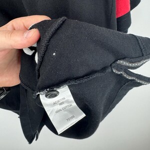 Może przedstawiać: Zbliżenie czarnej koszulki polo z czerwonymi akcentami na rękawie. Etykieta głosi "AE90155 RED 100% COTTON 70169". Koszulka wykonana jest z miękkiego, wygodnego materiału.