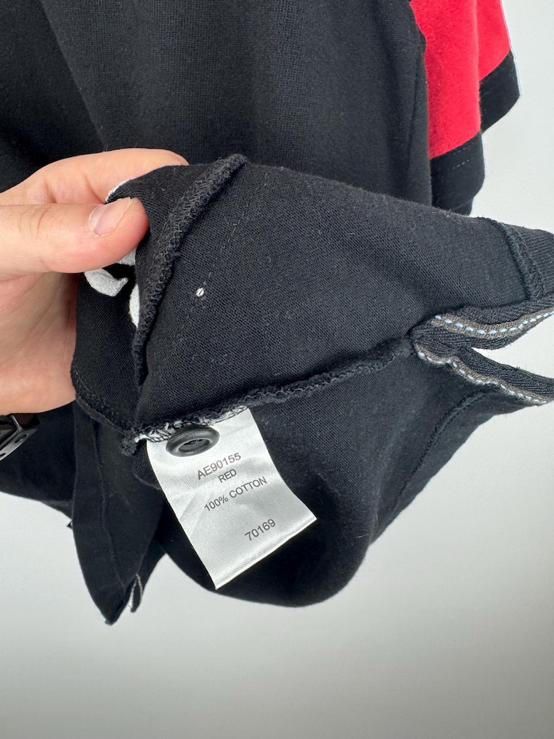 Może przedstawiać: Zbliżenie czarnej koszulki polo z czerwonymi akcentami na rękawie. Etykieta głosi "AE90155 RED 100% COTTON 70169". Koszulka wykonana jest z miękkiego, wygodnego materiału.