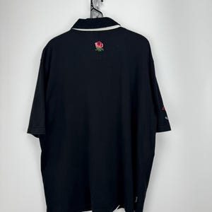 Puede incluir: Polo negro con cuello blanco y una rosa roja bordada en la espalda. La camisa tiene mangas cortas y un peque&ntilde;o logo en la manga. La camisa est&aacute; colgada de una cadena de metal.