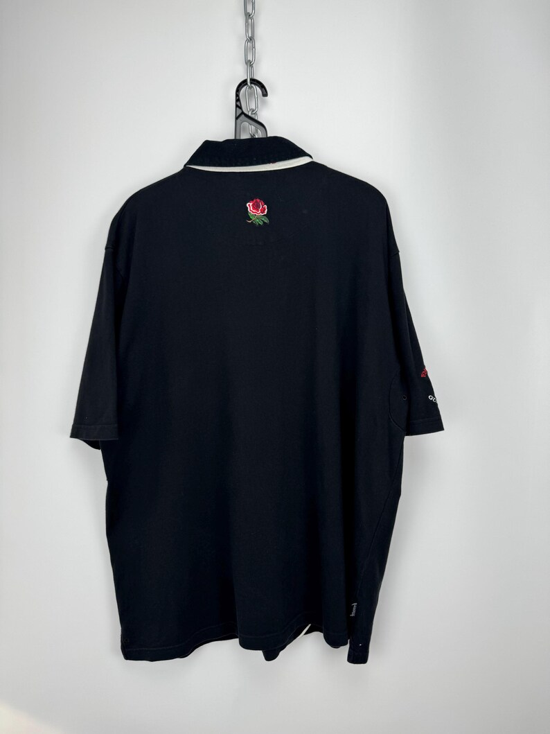 Puede incluir: Polo negro con cuello blanco y una rosa roja bordada en la espalda. La camisa tiene mangas cortas y un peque&ntilde;o logo en la manga. La camisa est&aacute; colgada de una cadena de metal.
