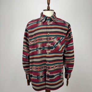 Puede incluir: Una camisa abotonada con un estampado geométrico colorido en rojo, beige, turquesa y naranja. La camisa tiene un bolsillo en el pecho y se exhibe en un maniquí de madera.