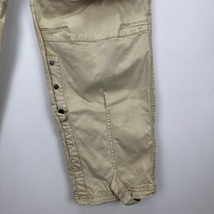 May include: Khaki-colored cargo pants with multiple pockets and button details. The pants have a relaxed fit and are made of a durable fabric. The design includes reinforced stitching and a strap detail at the hem.