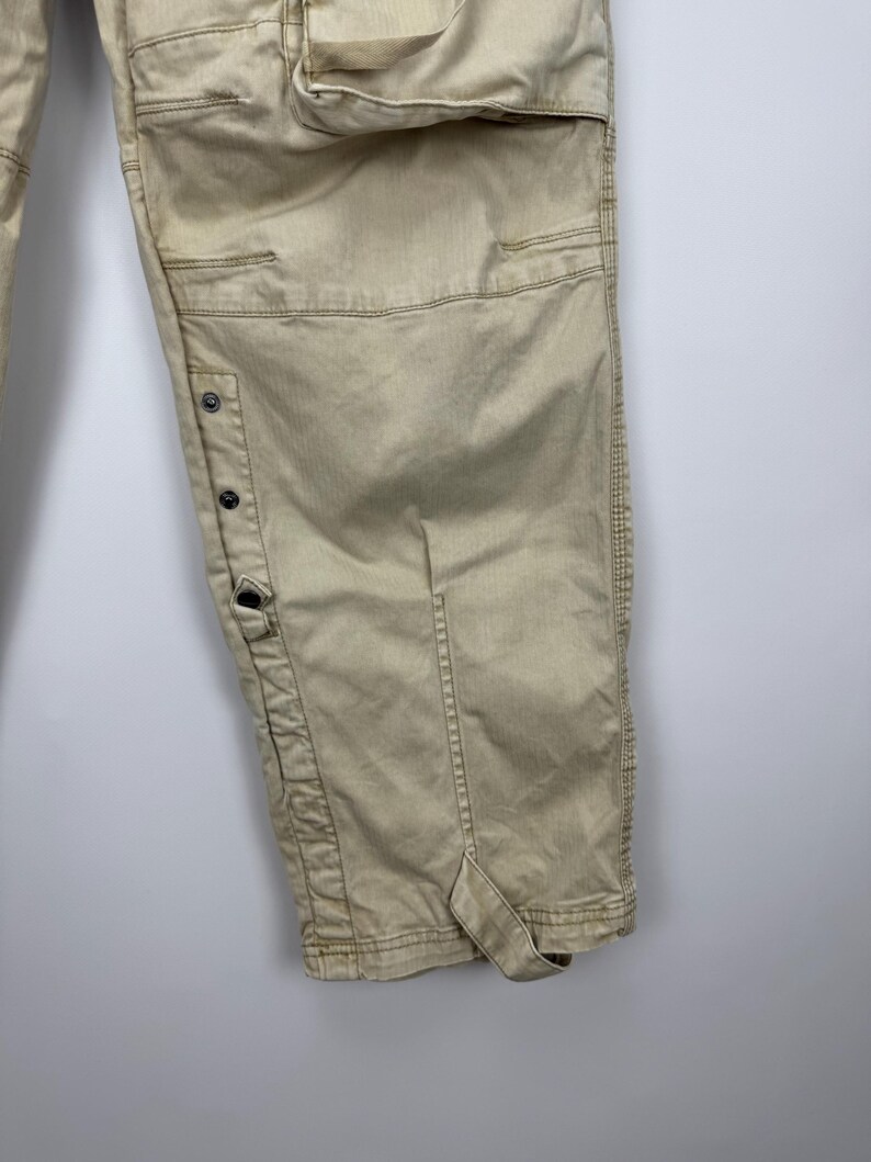 May include: Khaki-colored cargo pants with multiple pockets and button details. The pants have a relaxed fit and are made of a durable fabric. The design includes reinforced stitching and a strap detail at the hem.