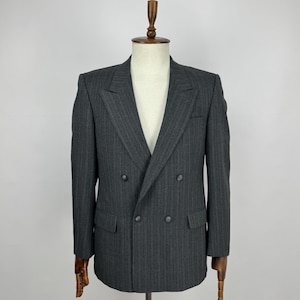 Puede incluir: Un blazer cruzado gris carbón con un sutil patrón de rayas finas. El blazer presenta solapas con muescas, dos filas de botones y dos bolsillos delanteros. La chaqueta se exhibe en un maniquí de madera.