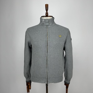 Może przedstawiać: Szary rozpinany sweter z żebrowanym wzorem. Sweter ma wysoki kołnierz zapinany na guzik i małą haftowaną naszywkę na piersi. Rękawy mają paski na mankietach. Sweter jest prezentowany na drewnianym manekinie. Wymiary w centymetrach.