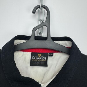 Puede incluir: Polo negro con cuello blanco y ribete rojo. La camisa presenta un parche con el logotipo de Guinness y el texto "ESTD 1759 GUINNESS OFFICIAL MERCHANDISE". La camisa est&aacute; colgada de una percha negra sujeta a una cadena plateada.