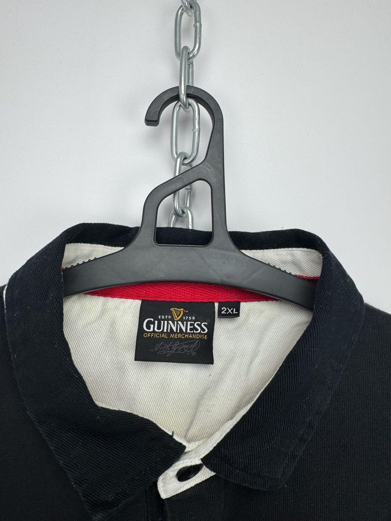 Puede incluir: Polo negro con cuello blanco y ribete rojo. La camisa presenta un parche con el logotipo de Guinness y el texto "ESTD 1759 GUINNESS OFFICIAL MERCHANDISE". La camisa est&aacute; colgada de una percha negra sujeta a una cadena plateada.