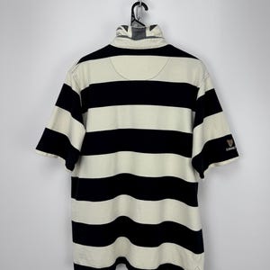 Puede incluir: Polo a rayas crema y azul marino de manga corta. La camisa tiene cuello y un peque&ntilde;o logotipo de Guinness en la manga. Cuelga de una percha negra.