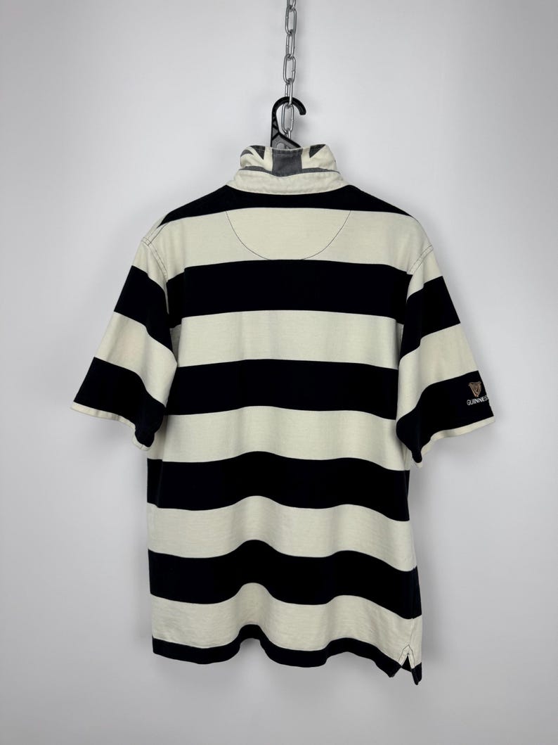 Puede incluir: Polo a rayas crema y azul marino de manga corta. La camisa tiene cuello y un peque&ntilde;o logotipo de Guinness en la manga. Cuelga de una percha negra.