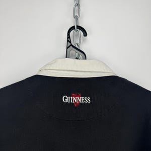 Puede incluir: Camisa negra de manga larga con cuello color crema. La camisa presenta el logotipo de Guinness bordado en rojo y blanco en el pecho. La camisa est&aacute; colgada de una percha negra sujeta a una cadena plateada.