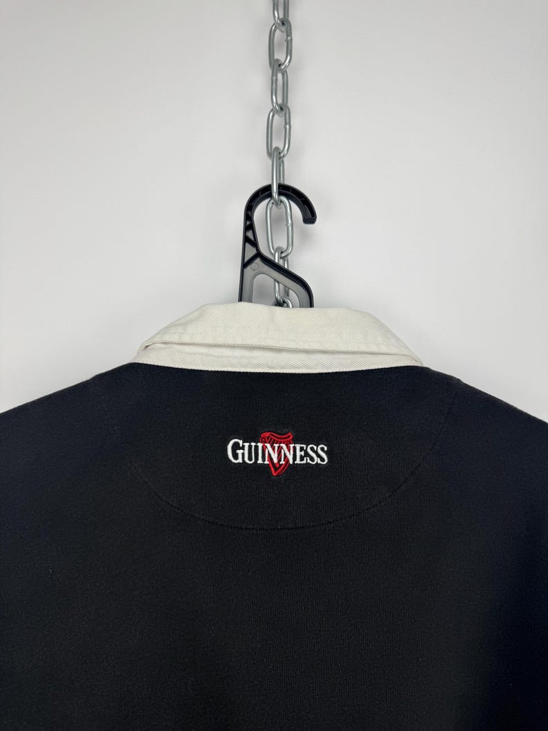 Puede incluir: Camisa negra de manga larga con cuello color crema. La camisa presenta el logotipo de Guinness bordado en rojo y blanco en el pecho. La camisa est&aacute; colgada de una percha negra sujeta a una cadena plateada.
