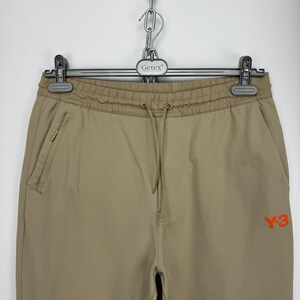 May include: Tan athletic pants with a drawstring waist and side pockets. The pants feature an orange "Y-3" logo on the right leg. The pants are hanging from a metal hanger.