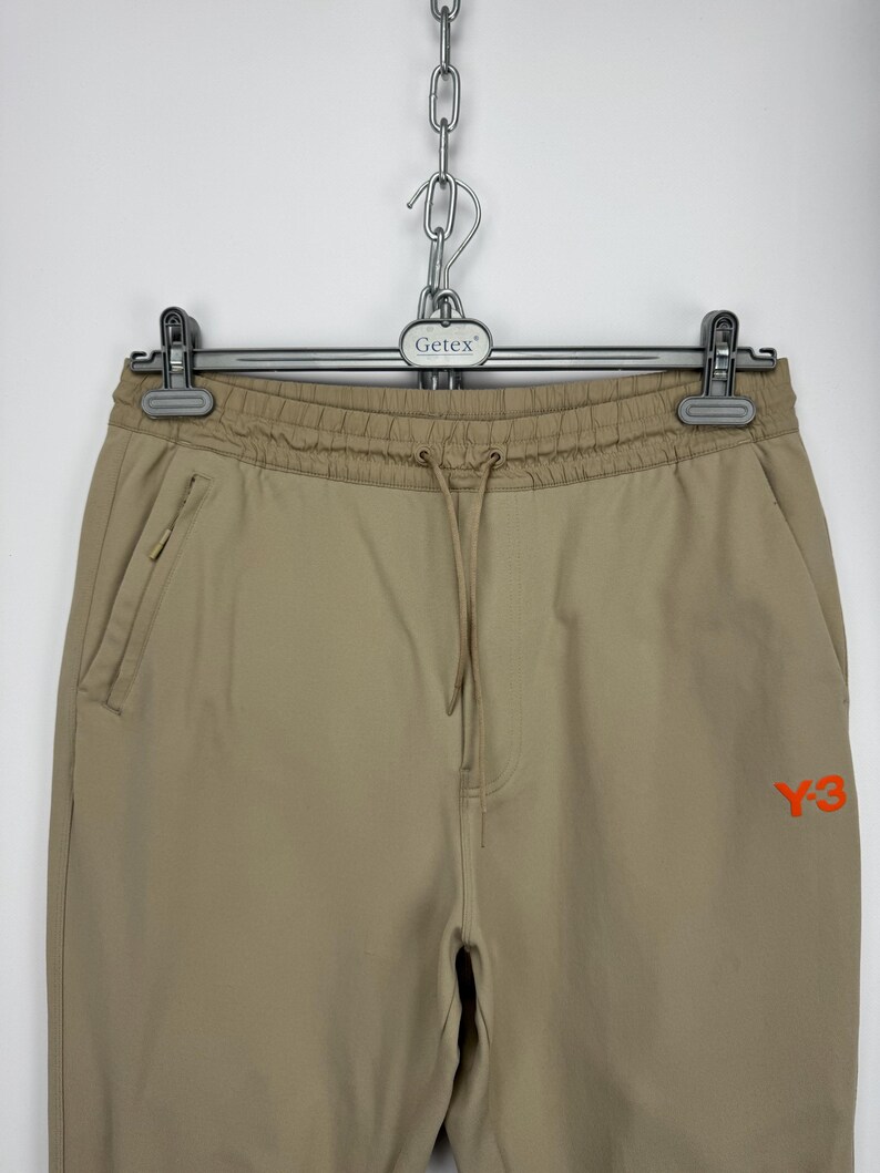 May include: Tan athletic pants with a drawstring waist and side pockets. The pants feature an orange "Y-3" logo on the right leg. The pants are hanging from a metal hanger.