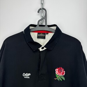 Puede incluir: Camiseta de rugby negra con cuello blanco y bordado de rosa roja. Presenta el texto "Cotton Traders" y "Guinness" en el cuello. Tiene una tapeta con botones y mangas largas. Una prenda superior cl&aacute;sica e informal.