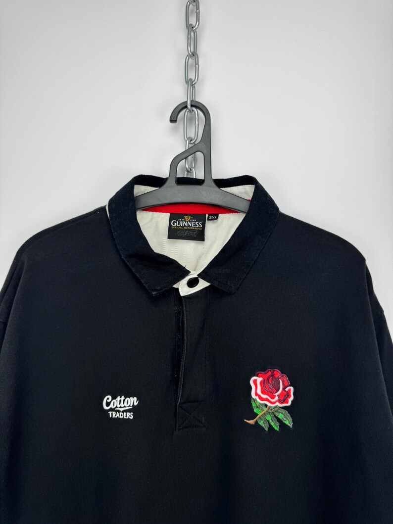 Puede incluir: Camiseta de rugby negra con cuello blanco y bordado de rosa roja. Presenta el texto "Cotton Traders" y "Guinness" en el cuello. Tiene una tapeta con botones y mangas largas. Una prenda superior cl&aacute;sica e informal.