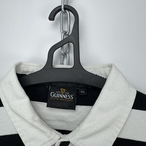 May include: A black and white striped polo shirt with a white collar and the Guinness logo. The shirt is hanging on a black hanger. The shirt is labelled XL.