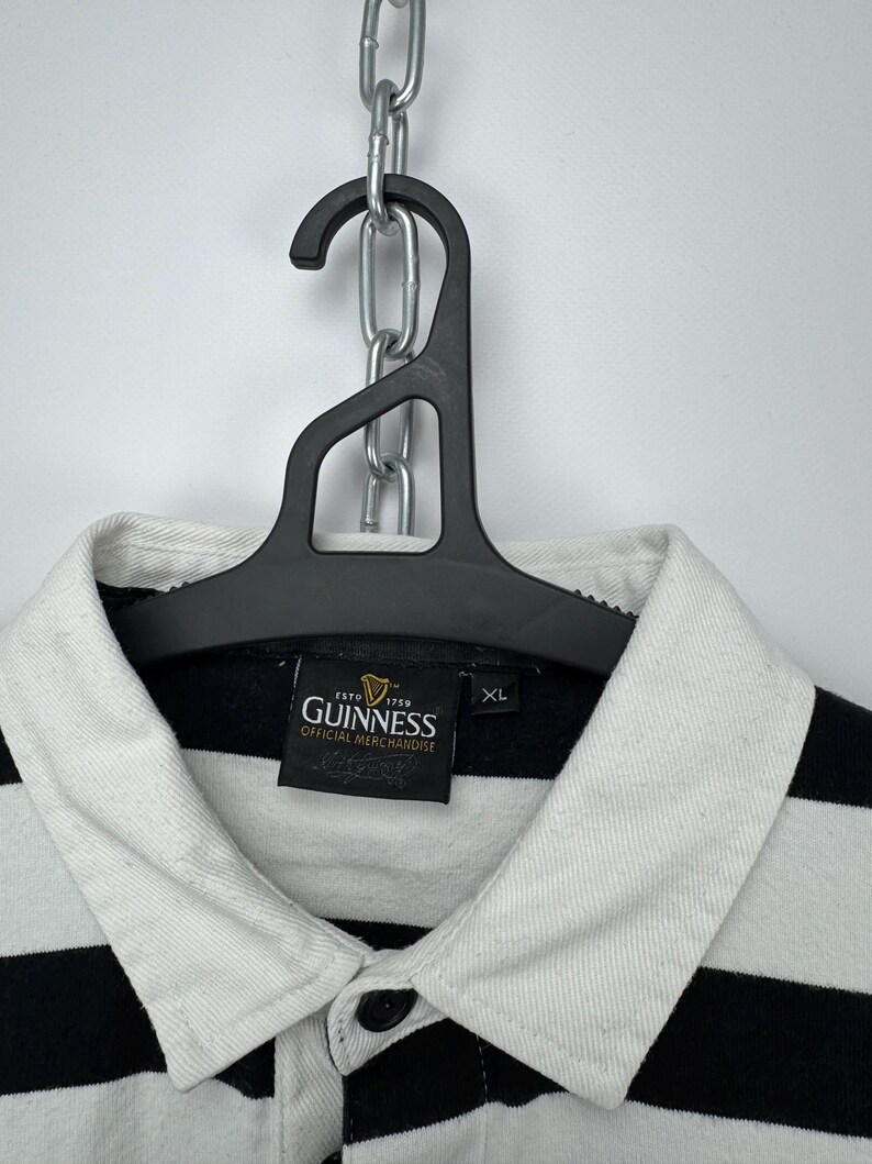 May include: A black and white striped polo shirt with a white collar and the Guinness logo. The shirt is hanging on a black hanger. The shirt is labelled XL.