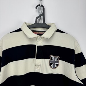 Puede incluir: Polo a rayas crema y azul marino con cuello cl&aacute;sico y tapeta de tres botones. Cuenta con un parche de escudo con un dise&ntilde;o de la Union Jack en el pecho. La camisa est&aacute; colgada en una percha negra.