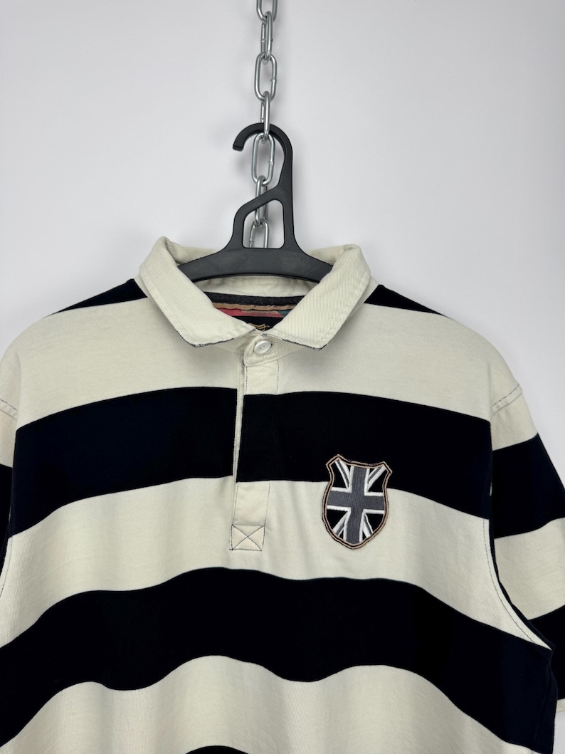 Puede incluir: Polo a rayas crema y azul marino con cuello cl&aacute;sico y tapeta de tres botones. Cuenta con un parche de escudo con un dise&ntilde;o de la Union Jack en el pecho. La camisa est&aacute; colgada en una percha negra.