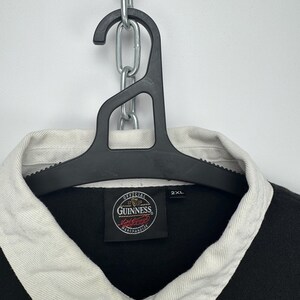Puede incluir: Camisa de rugby negra con cuello blanco y tapeta abotonada. La camisa presenta el logotipo de Guinness y la talla 2XL. La camisa est&aacute; colgada de una percha negra.