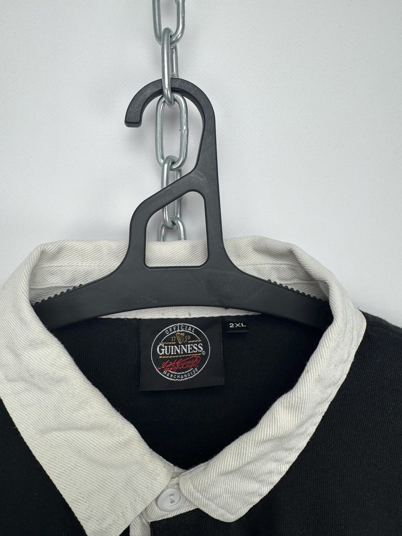 Puede incluir: Camisa de rugby negra con cuello blanco y tapeta abotonada. La camisa presenta el logotipo de Guinness y la talla 2XL. La camisa est&aacute; colgada de una percha negra.