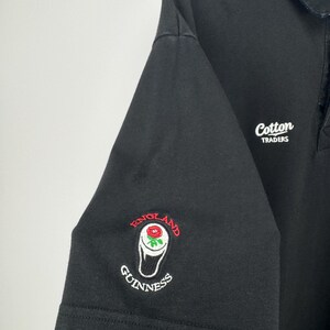 Puede incluir: Polo negro con cuello y ribetes de manga blancos. Presenta el texto bordado "Cotton Traders" y un logotipo de Guinness con una rosa y las palabras "England" y "Guinness".