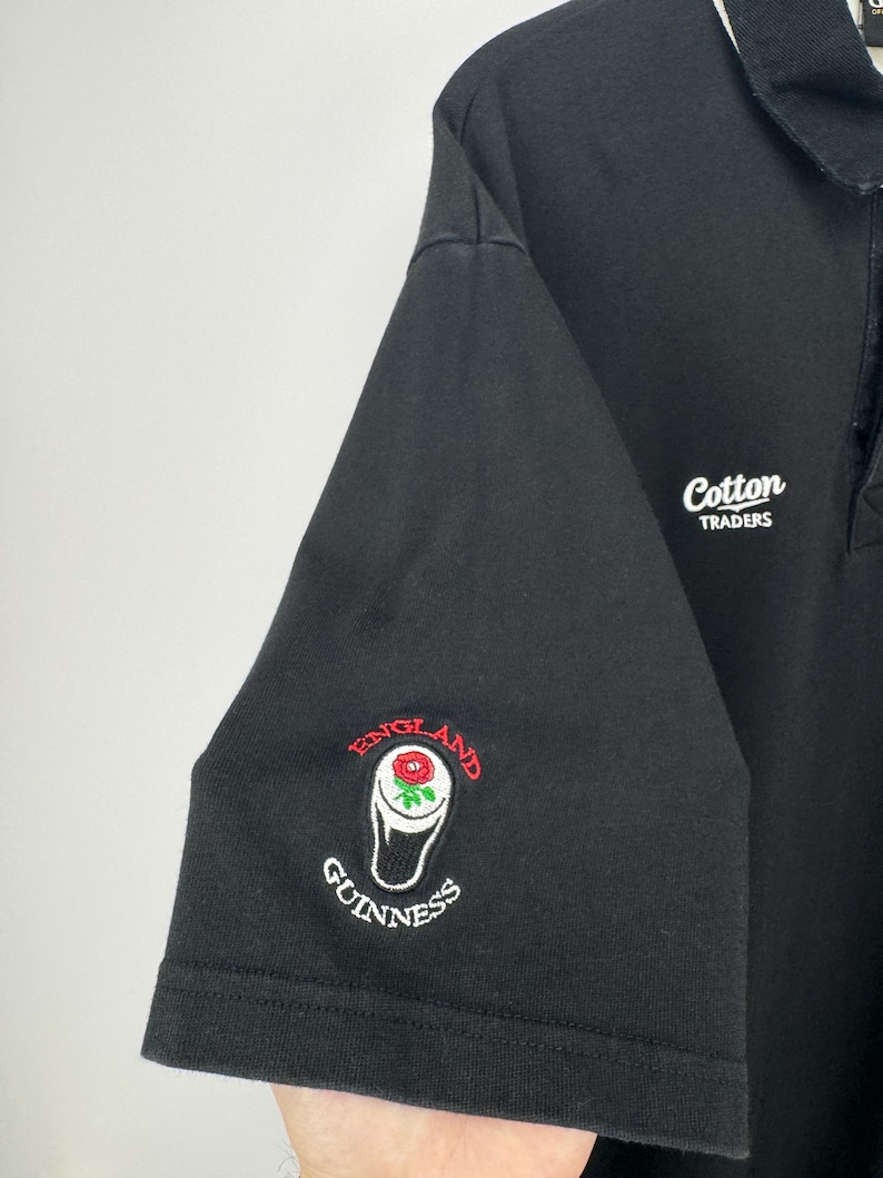 Puede incluir: Polo negro con cuello y ribetes de manga blancos. Presenta el texto bordado "Cotton Traders" y un logotipo de Guinness con una rosa y las palabras "England" y "Guinness".