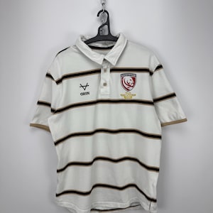 May include: White rugby shirt with horizontal black and tan stripes. Features a collared neckline, short sleeves, and a three-button placket. The shirt has a team logo and the text "Gloucester Rugby" on the chest, with the Oxen logo.