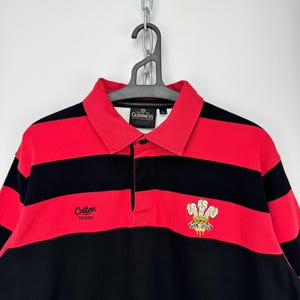 Może przedstawiać: Koszulka do rugby w czerwono-czarne poziome paski. Koszulka ma kołnierz i napis "Cotton Traders" oraz herb. Koszulka wisi na czarnym wieszaku.