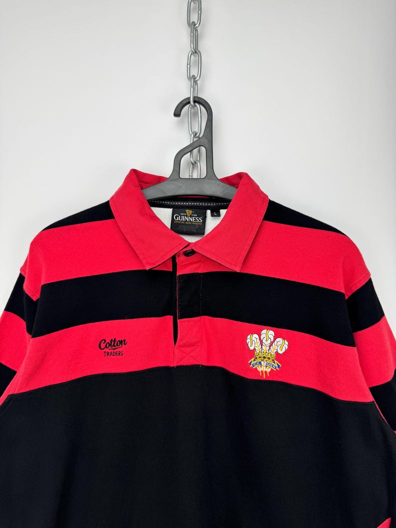 Może przedstawiać: Koszulka do rugby w czerwono-czarne poziome paski. Koszulka ma kołnierz i napis "Cotton Traders" oraz herb. Koszulka wisi na czarnym wieszaku.