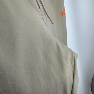 May include: Tan-colored athletic pants with a drawstring and a small orange logo on the upper right side. The pants are made of a soft, textured fabric and have a relaxed fit. The drawstring is a matching tan color.