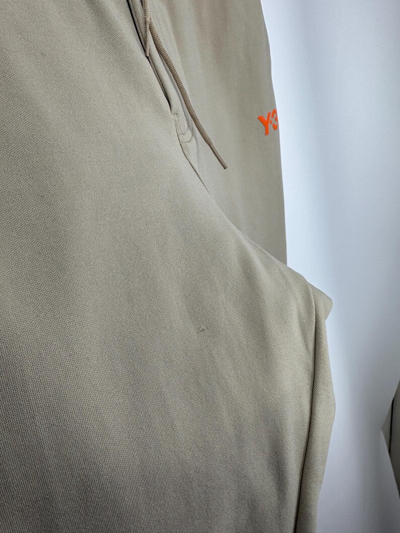 May include: Tan-colored athletic pants with a drawstring and a small orange logo on the upper right side. The pants are made of a soft, textured fabric and have a relaxed fit. The drawstring is a matching tan color.
