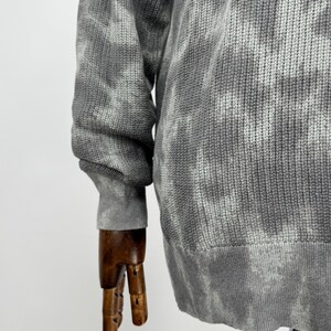 May include: A grey tie-dye patterned sweater. The long-sleeved sweater has a ribbed hem and cuffs. The fabric appears to be a knit material. The sweater is displayed on a wooden mannequin arm.