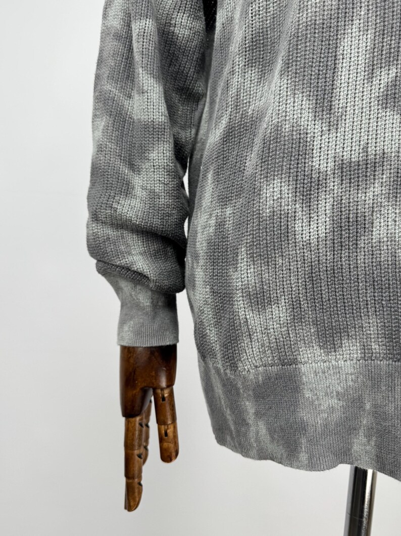 May include: A grey tie-dye patterned sweater. The long-sleeved sweater has a ribbed hem and cuffs. The fabric appears to be a knit material. The sweater is displayed on a wooden mannequin arm.