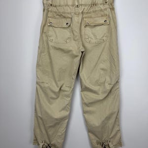 May include: Khaki-colored cropped pants with two back pockets and elastic waistband. The pants have a relaxed fit and feature tie details at the ankles. The garment is hanging on a hanger against a plain background.