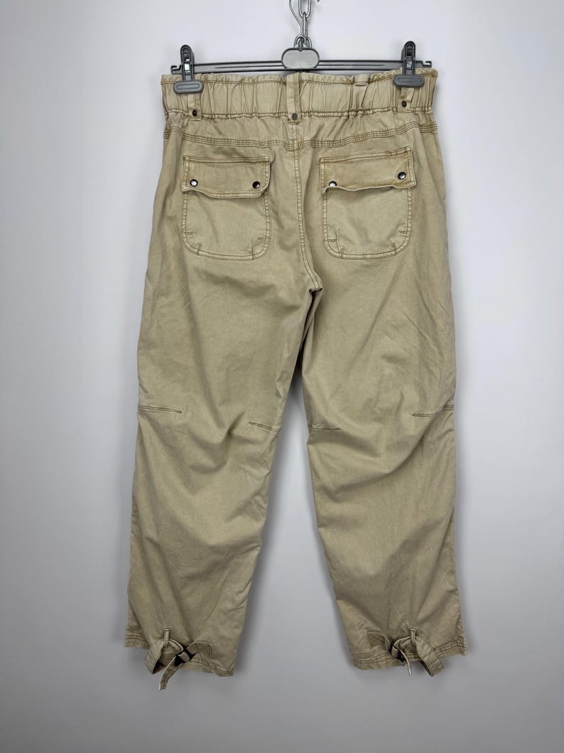 May include: Khaki-colored cropped pants with two back pockets and elastic waistband. The pants have a relaxed fit and feature tie details at the ankles. The garment is hanging on a hanger against a plain background.