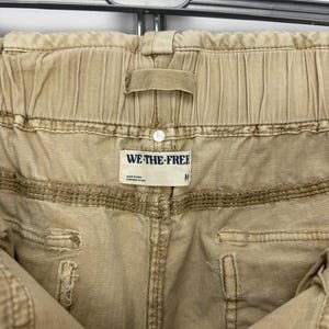 May include: Close-up of tan-colored pants with an elastic waistband. The pants have visible stitching and a small tag that reads "WE THE FREE" and the size "M". The pants are hanging on a metal hanger.