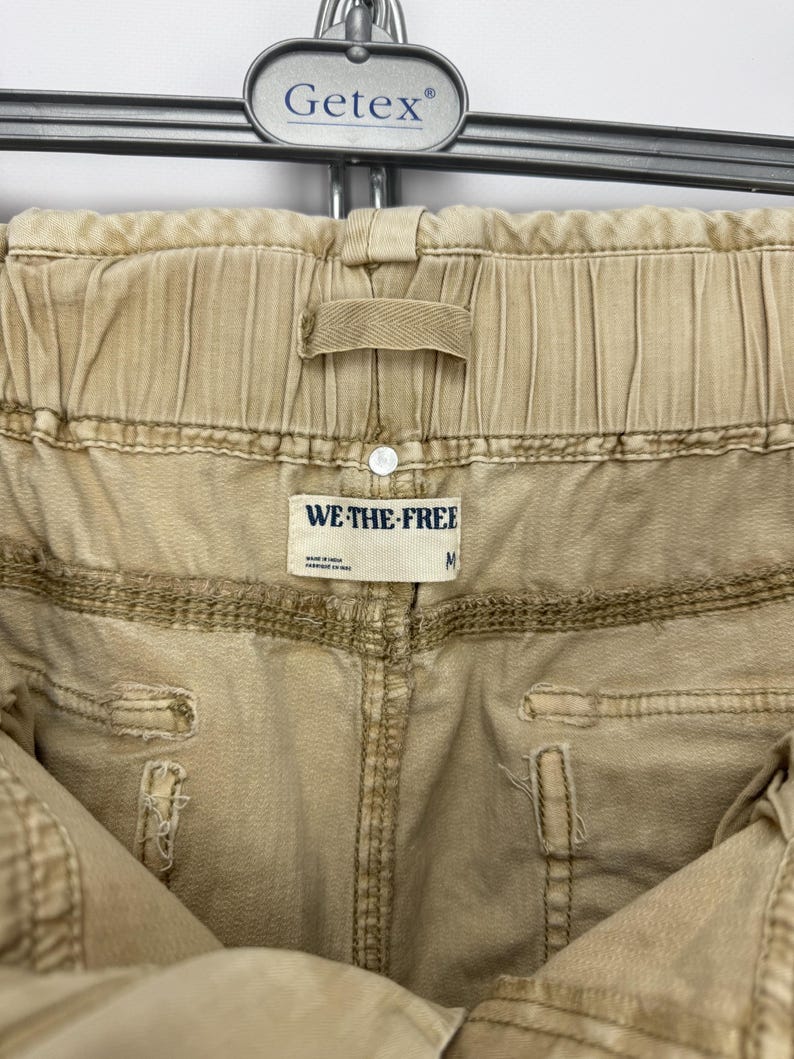 May include: Close-up of tan-colored pants with an elastic waistband. The pants have visible stitching and a small tag that reads "WE THE FREE" and the size "M". The pants are hanging on a metal hanger.